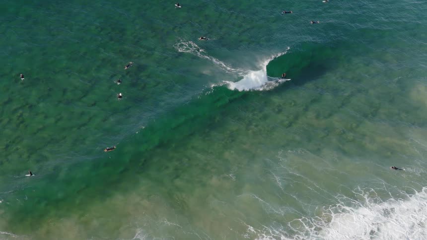 An aerial shot shows surfers waiting in the ocean as a large wave breaks and rushes toward the shore a person on a surfboard can be seen riding the wave as it crests and turns into white foam