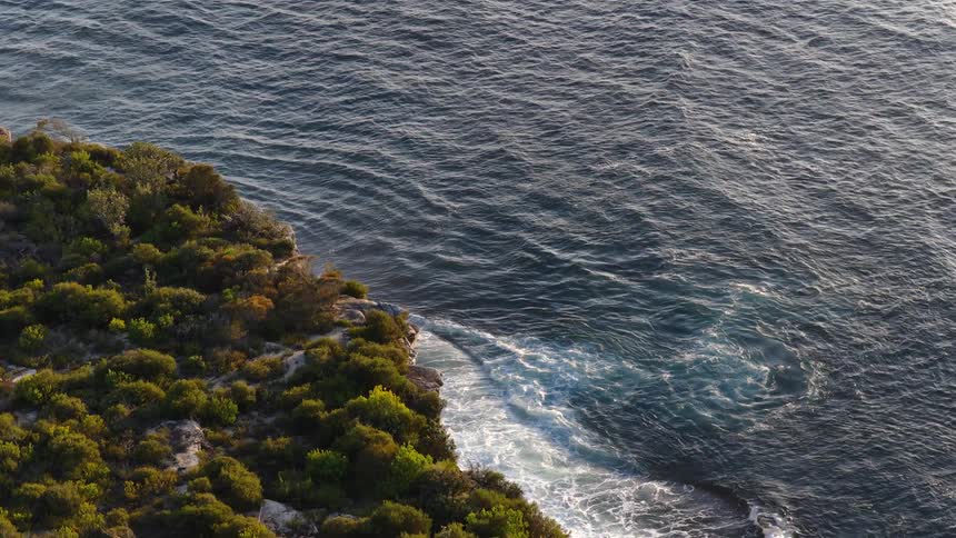 An aerial shot slowly pulls back from a rocky bushcovered coastline where the deep blue ocean waves are gently crashing against the cliffs