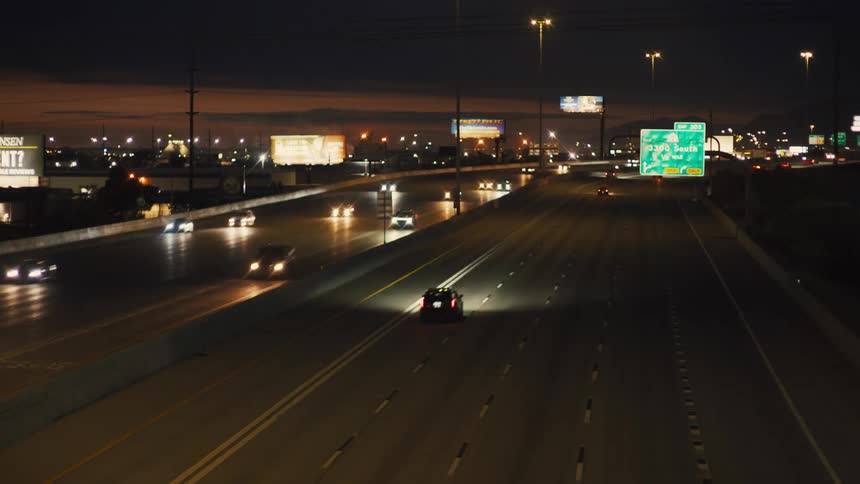 Cars drive on a highway lit at night with city in the background