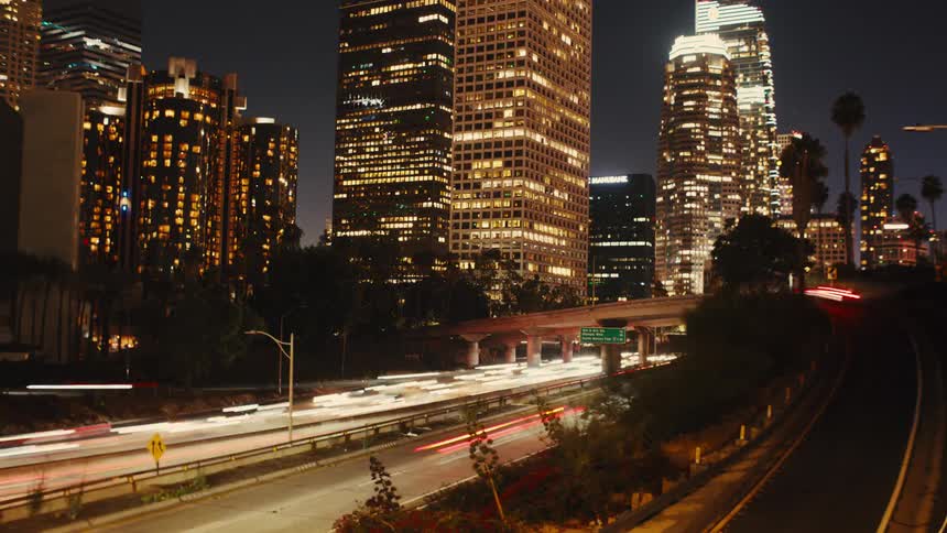 A nighttime long exposure view of a city shows a multilane highway in the foreground with blurred streaks of red and white lights from passing traffic tall buildings with illuminated windows in the background and palm trees visible between the highway