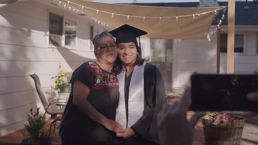 An elderly woman wearing glasses stands beside a young girl in a graduation gown both posing for a photo while someone takes a snapshot with a phone they smile as they stand on a front porch at midday