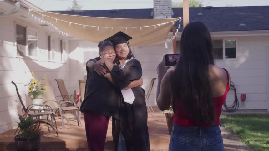Graduate in cap and gown hugs older woman while another takes a picture of the scene