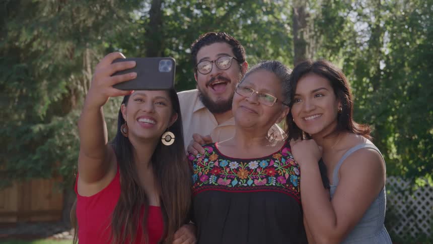 A family of fourtwo young sisters a brother and an elderly mothertaking a selfie with a phone in their yard all smiling at midday