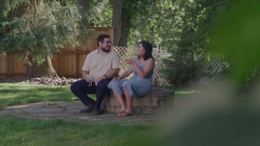 A couple is seen sitting on the edge of a circular stone fire pit in a lush green backyard engaged in a conversation on a sunny day