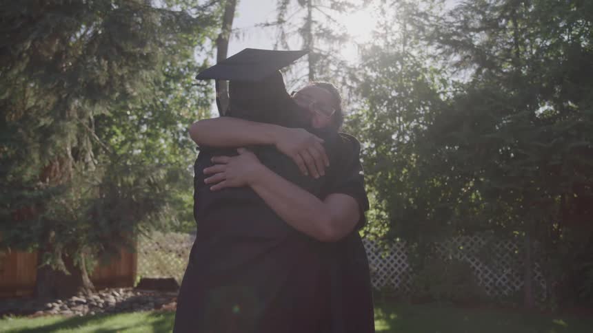 A woman wearing glasses and a dark shirt hugs a person in a graduation cap and gown while standing in a bright sunlit backyard with trees and a lattice fence
