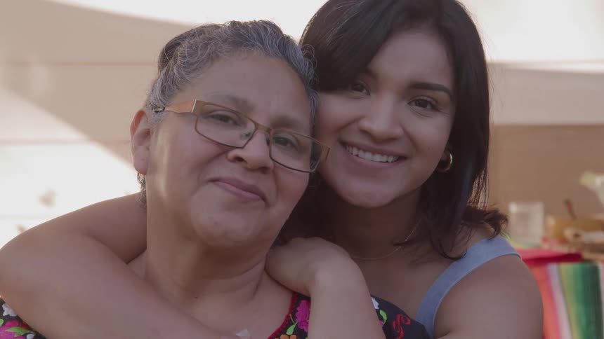 A younger woman with brown hair embraces an older woman with gray hair and glasses from behind resting her cheek on the older womans head and both women look forward smiling at the camera