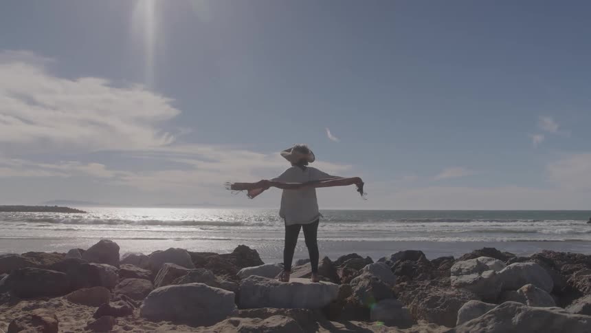 A young woman in a sun hat is standing on some rocks near the ocean she holds her arms out and raises a shawl above her head