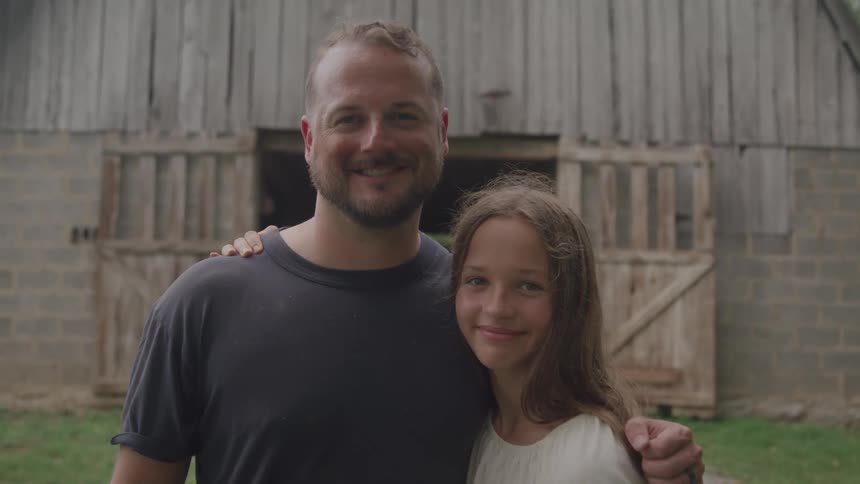 A father with a beard and a dark gray tshirt stands with his arm around his daughter who has long brown hair and a white top as they both smile at the camera in front of a rustic wooden barn