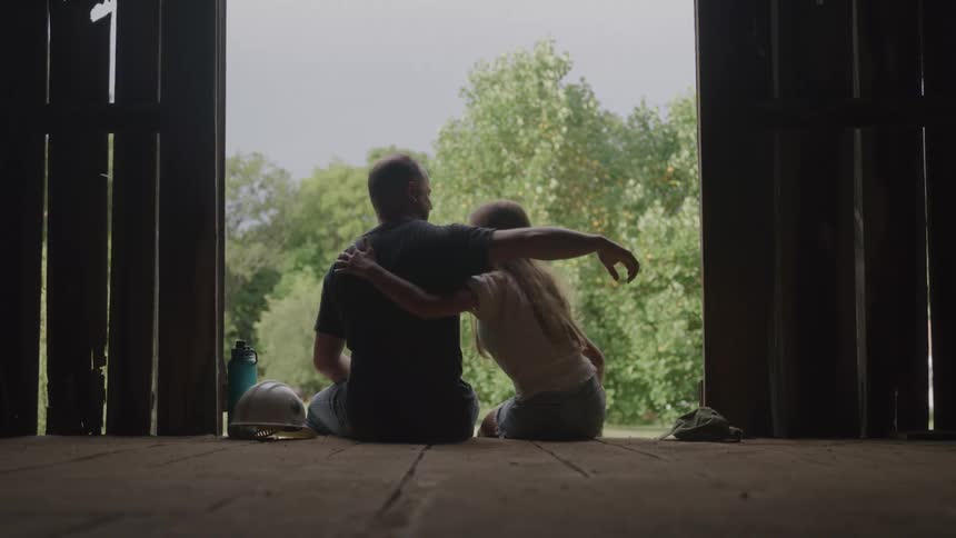 Man and a child sitting in a barn hugging while watching nature