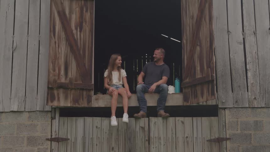 A father and his young daughter sit on the open doorway of a large rustic barn looking at each other and smiling