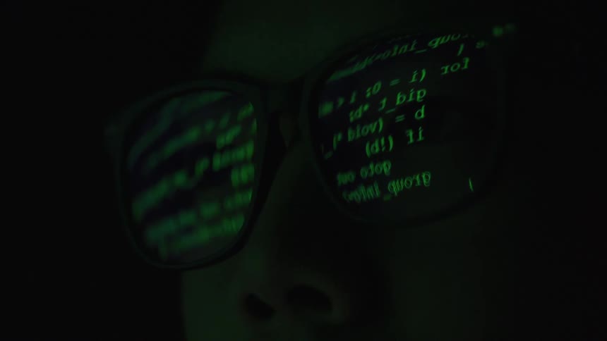 A video shows a closeup of a person wearing glasses with green computer code reflected in them the code scrolls and blurs as it moves
