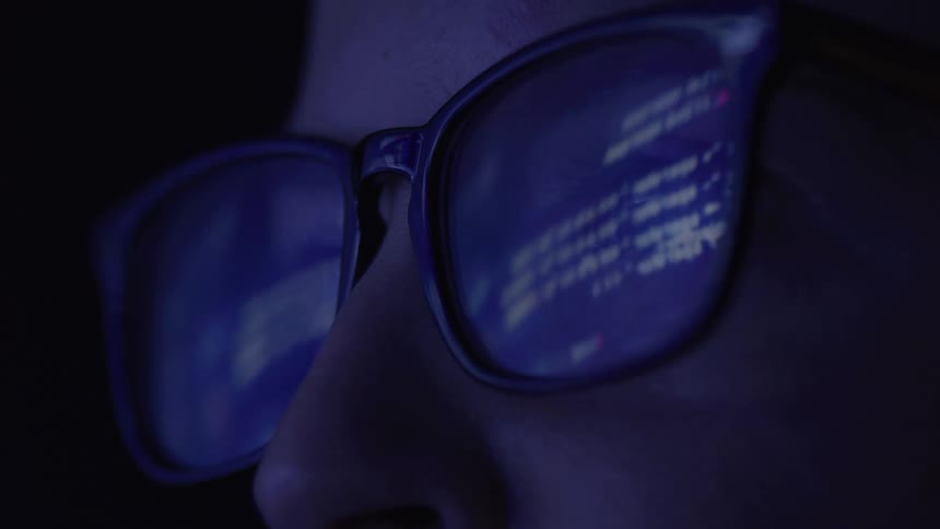 A closeup shot shows a persons face in a dark room the light from a computer screen is reflected in their glasses showing lines of code or data on the lenses
