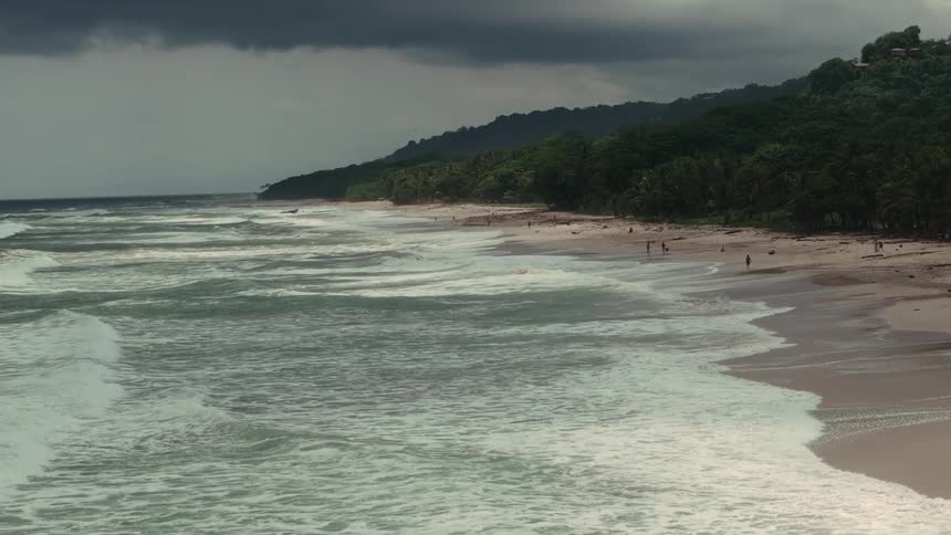 Strong waves crash onto a beach surrounded by forest under a cloudy sky