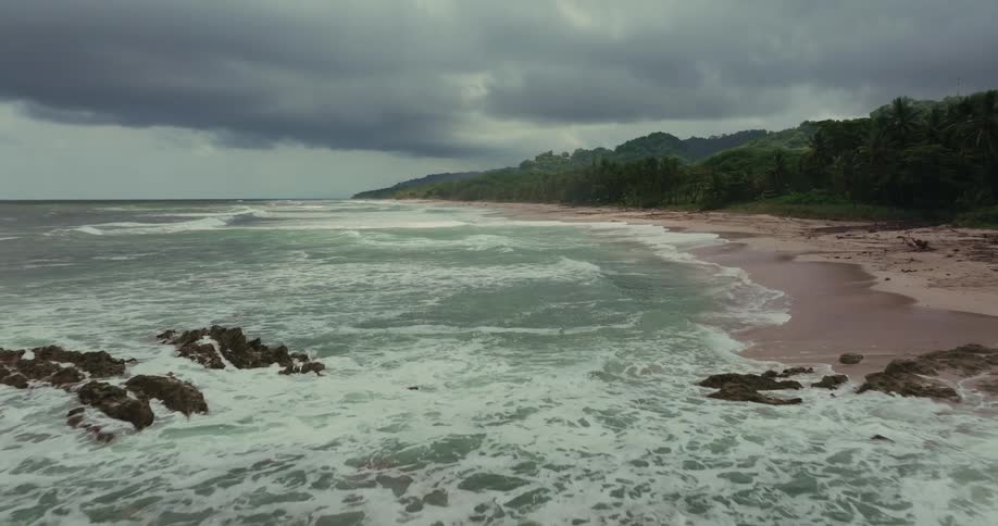 A long sandy beach with a dense green forest on the far side all under a dark cloudy sky