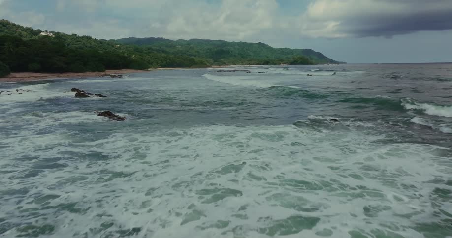 Waves crash against rocks on a beach surrounded by green hills
