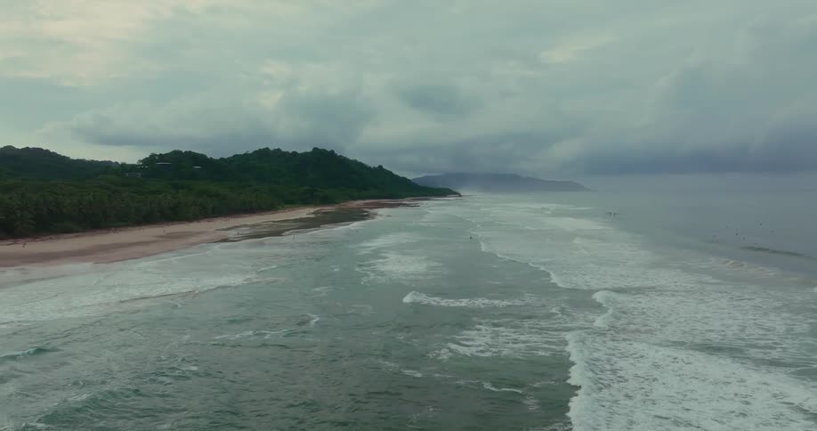 An aerial shot shows ocean waves approaching a wide sandy beach the coastline is bordered by a lush green mountain range that extends into the distance