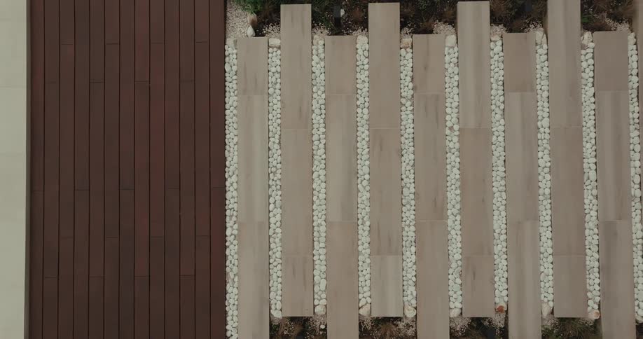 An overhead shot shows a modern patio featuring a pool on the left and a pathway made of alternating wooden planks and strips of river rocks on the right