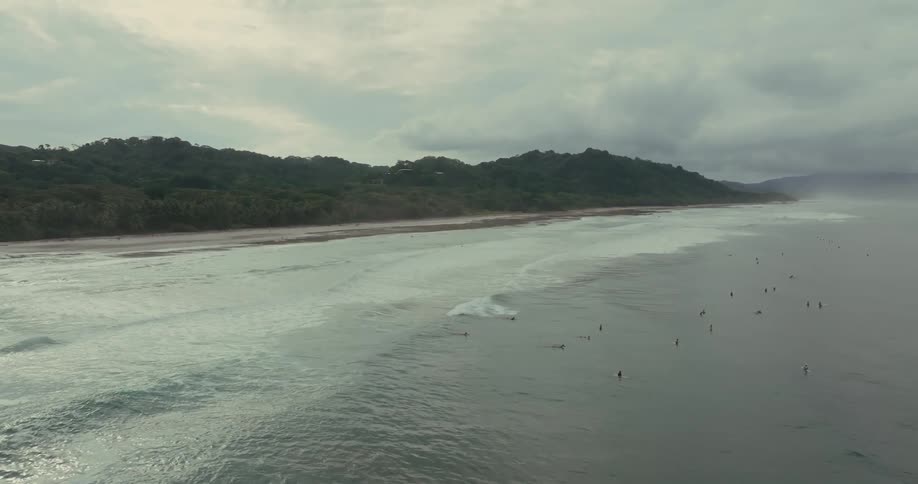 Group of surfers waits for waves in the sea on a cloudy day near the green coast