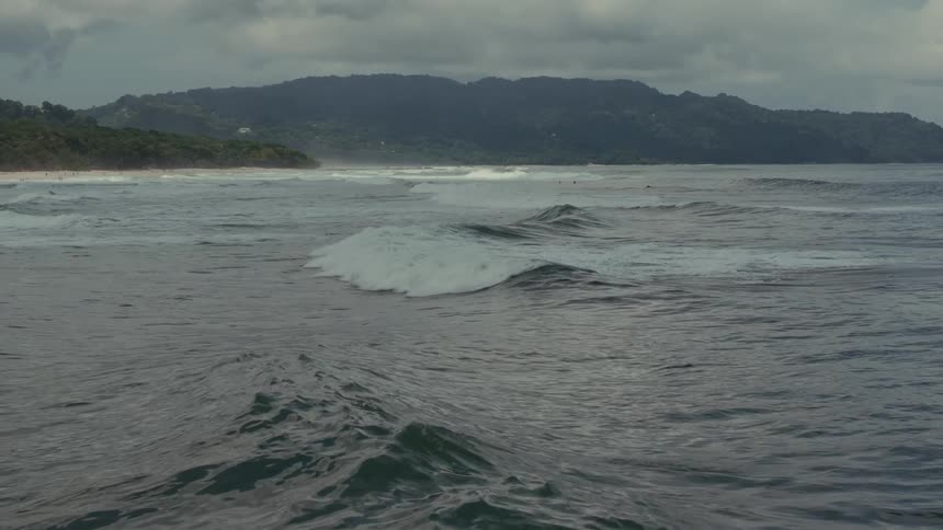 An aerial shot captures large waves on a gray overcast day the waves crash onto a beach at the foot of a dark forested mountain range