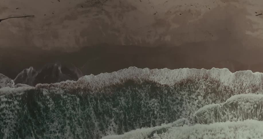 An aerial view of an ocean shows a dark brown sandy beach and a line of light green waves with white foam crests crashing parallel to the shoreline