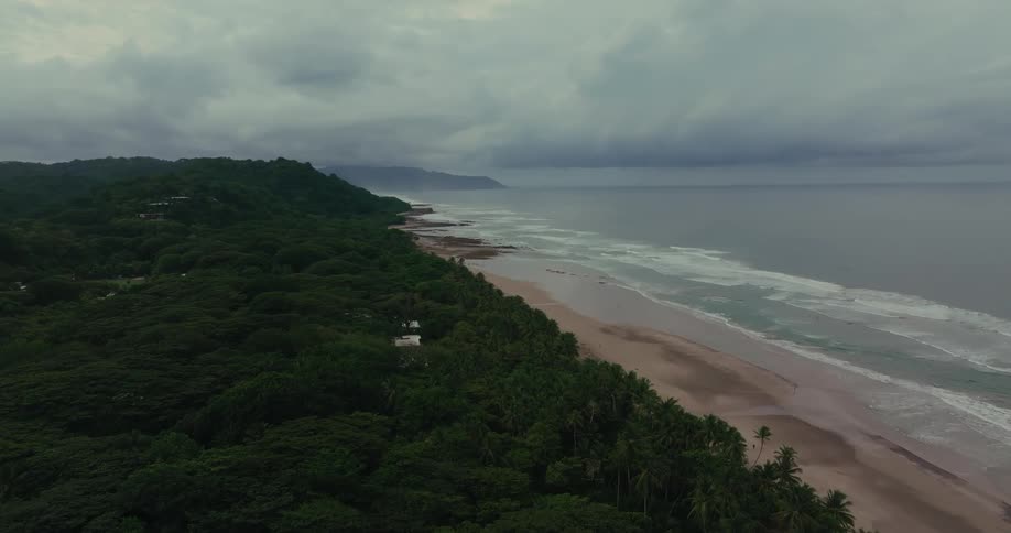 A slow aerial shot flies along a remote tropical coastline on a cloudy day showing a dense jungle meeting a wide sandy beach beside the ocean