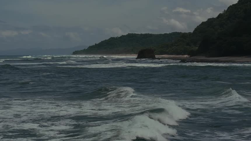 Rough waves approach the shore surrounded by green hills