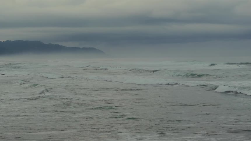 Waves crash on the ocean under a cloudy sky as birds fly above