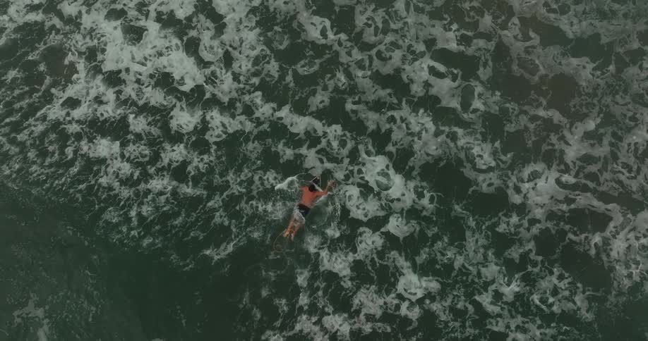 Two men wearing dark swim trunks and no shirts paddle on surfboards with one catching a large wave that carries him to the left as the other continues to paddle