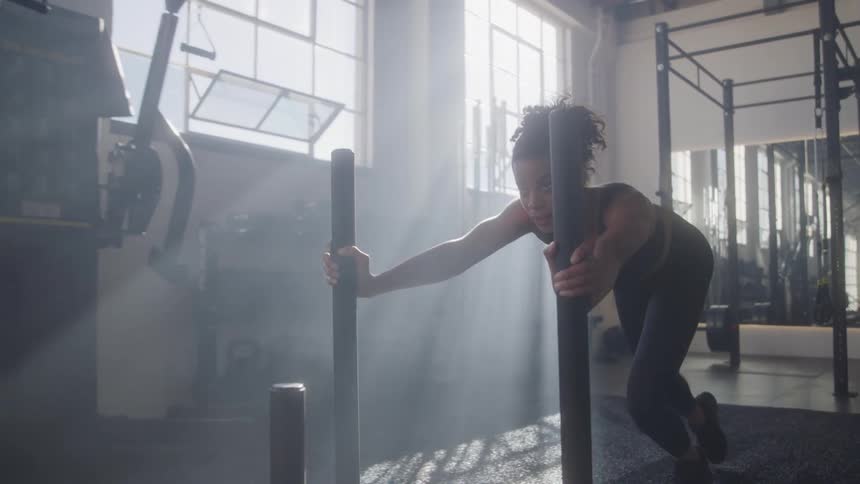 A woman is doing an athletic exercise in a gym with large windows she is pushing a weighted sled with both hands with one foot placed behind her and the other in front