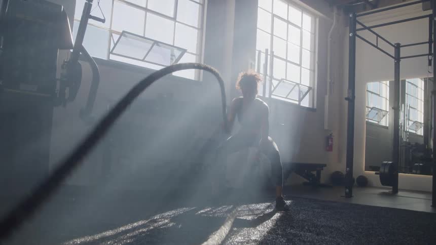 A woman forcefully whips heavy ropes up and down in a gym