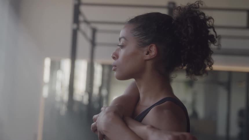 A woman with curly brown hair in a high ponytail is standing in a gym wearing a black top she is seen from the side with her body angled toward the right of the screen as she holds one arm across her chest to stretch her shoulder