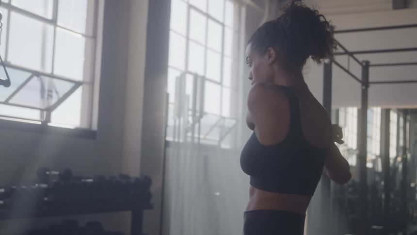 The video shows a side profile shot of a woman with dark curly hair wearing a black athletic top she is inside a gym with large windows stretching her shoulders and preparing to exercise