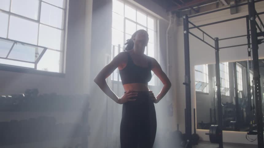 A woman with her hair in a bun stands in a dimly lit smoky gym wearing a black sports bra and pants with her hands on her hips as she takes a deep breath and looks up