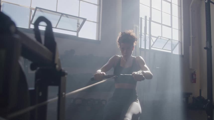 A woman is using a rowing machine in a welllit gym she is dressed in athletic wear and is actively pulling the handle toward her chest engaged in a workout