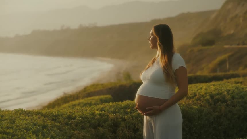 A pregnant woman holds her belly while looking out at the ocean