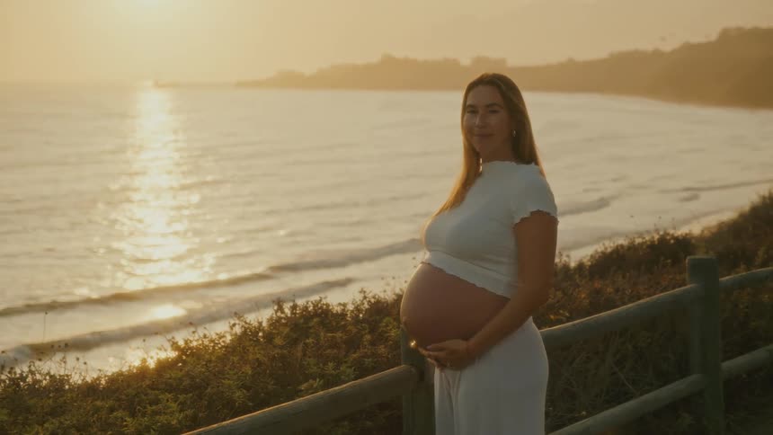 Pregnant woman smiles at sunset by the sea with hands on her belly