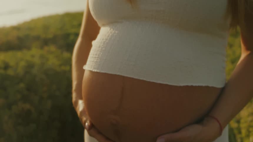 A smiling pregnant woman with long brown hair stands outdoors at dusk holding her bare stomach
