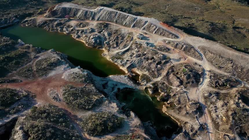 An aerial shot of a quarry shows two dark green bodies of water surrounded by brown dirt roads piles of dirt and gray rocks