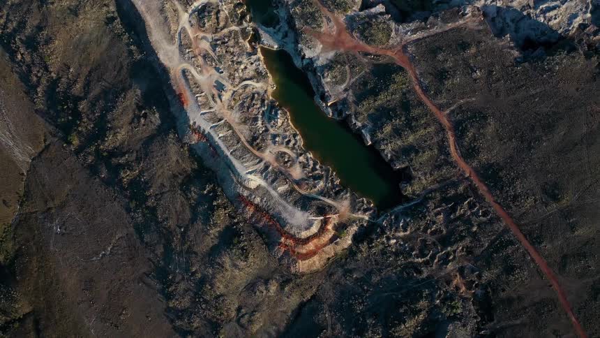 An aerial view of an openpit mine is shown with a long greentinted lake at the bottom of the crater the surrounding landscape is barren and dark with winding dirt roads leading to and from the site