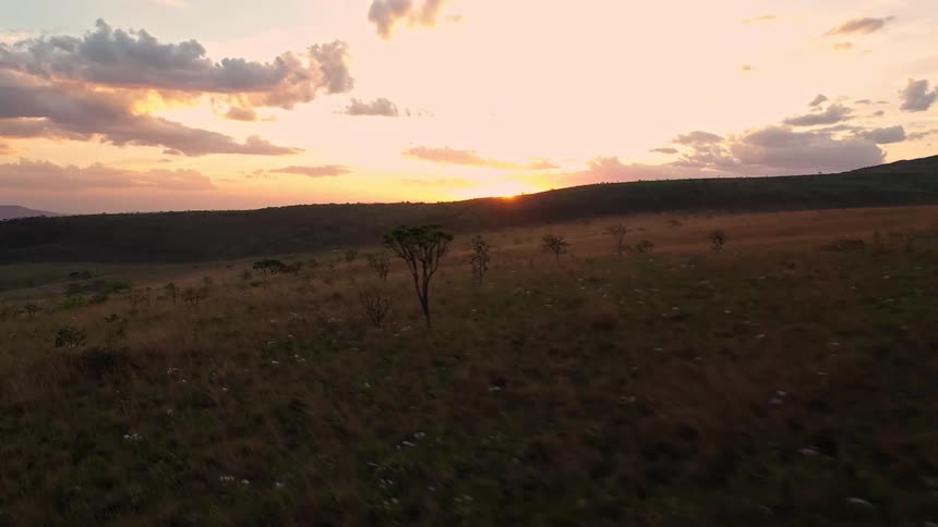 An aerial view shows a vast grassy field with a few scattered trees in the background the sun sets over a distant mountain range casting a warm glow across the sky