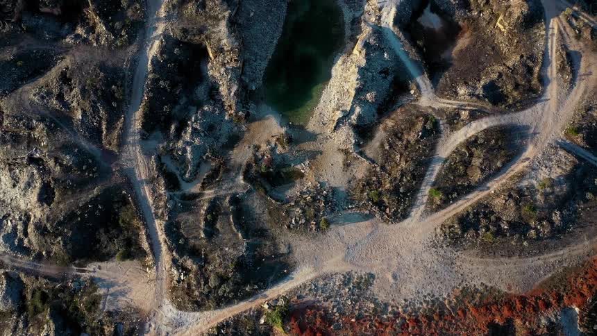 A topdown aerial shot slowly descends over a vast rocky quarry revealing a network of dirt roads and a small pool of water below