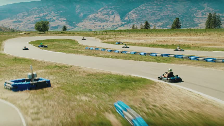 Several gokarts race around a paved track with blue and red barriers they speed past grassy areas and mountains with the camera capturing a wide shot of the action