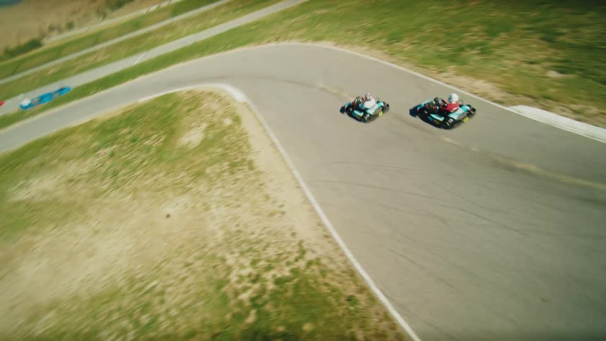 Two race car drivers wearing helmets and racing suits drive go karts on a race track with grassy fields on either side