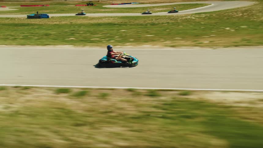A group of people in gokarts is racing around a paved track with one of the karts driving ahead of the others the track has green grass and hills on either side