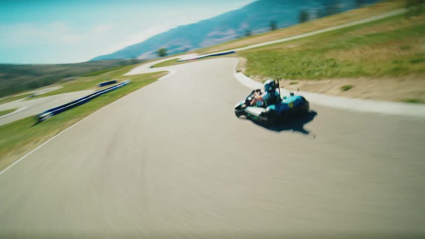 A drone follows a single go kart on a race track with several turns as a driver wearing a white helmet and blue shirt navigates the turns with a hilly mountainous landscape in the background