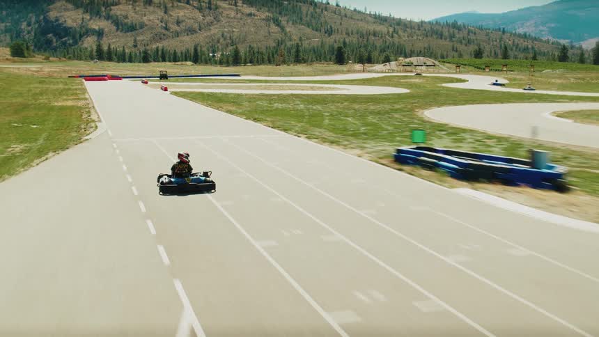 A wide aerial view shows gokarts racing on a winding track with a mountainous backdrop the camera follows the gokarts as they speed around the course