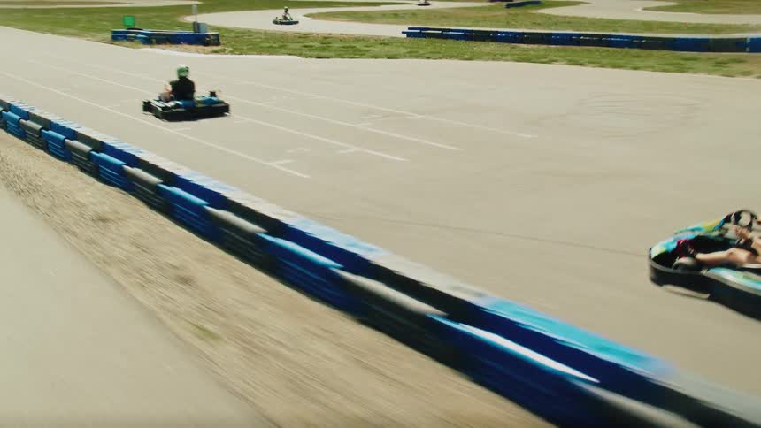 A view from the back of a go kart shows several drivers racing around a track on a sunny day with green hills and mountains in the distance