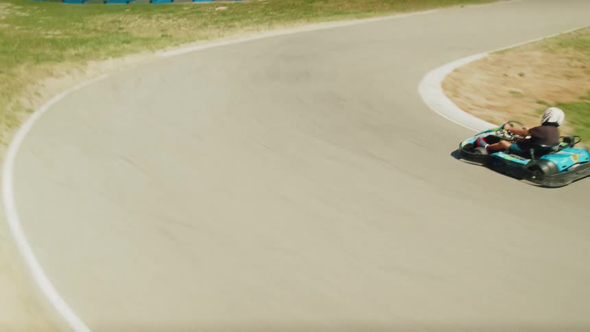 Several gokarts race around a paved track with blue and red barriers the track is set in a rural scenic area with mountains and green grass and hills in the background