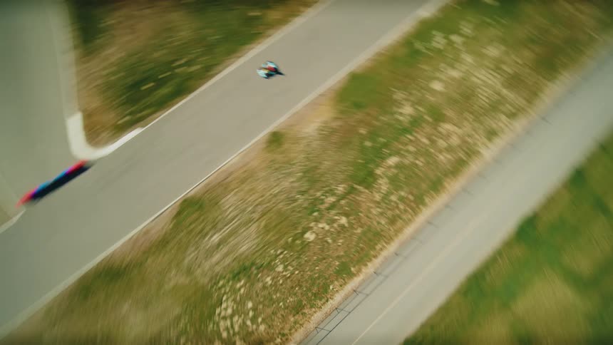 A drone follows a single go kart on a race track with several turns as a driver wearing a white helmet and red shirt navigates the turns with a hilly mountainous landscape in the background