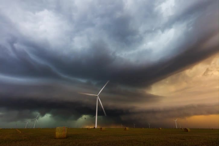 A dramatic storm cloud hangs low over a field with a wind turbine standing in the foreground the sky behind the clouds glows a vibrant orange contrasting with the dark storm